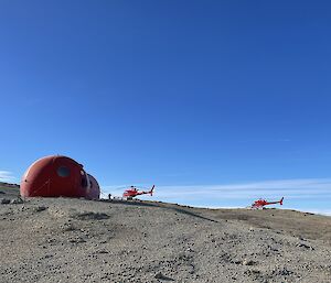 2 helicopters landing on dirt next to small round buildings