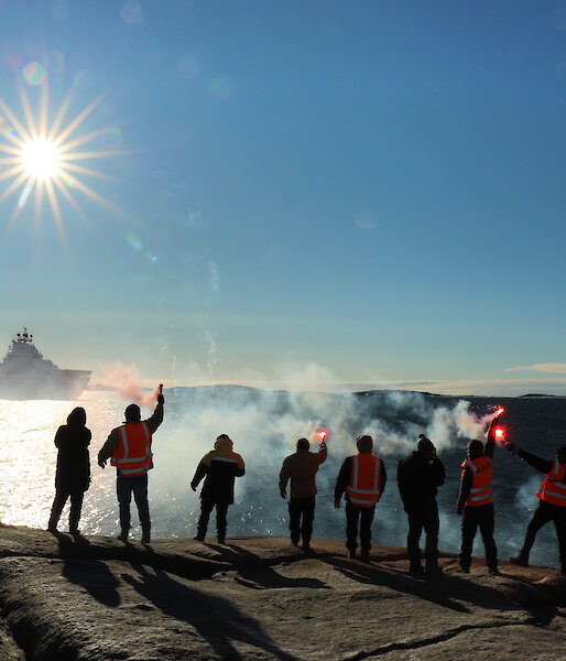 Ten people standing in a line on a rocky outcrop, with backs to camera and holding lit flares, wave farewell to a ship in the distance.