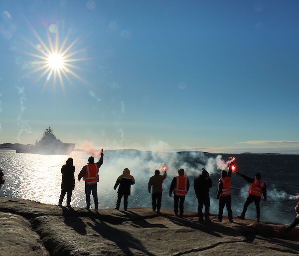 Ten people standing in a line on a rocky outcrop, with backs to camera and holding lit flares, wave farewell to a ship in the distance.