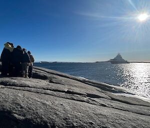 A group of people are huddled together on rocky outcrop at the front left, while distant right a ship is at anchor in the bay