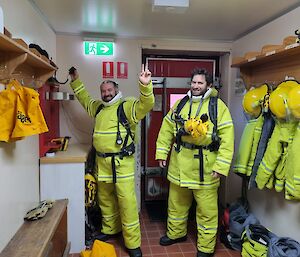 Two men wearing fire fighting uniforms and breathing aperatus on backs stand in porch surrounded by other fire fighting outfits hanging on hooks along the wall. Man on left raises both arms high into the air.