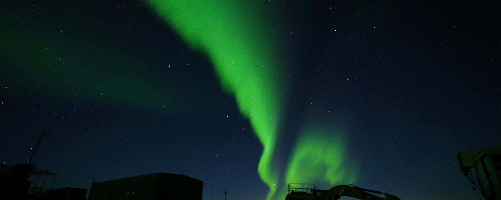 Green Aurora in thick line glows across centre of sky, in foreground in shadows are buildings and large machinery