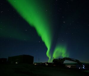 Green Aurora in thick line glows across centre of sky, in foreground in shadows are buildings and large machinery