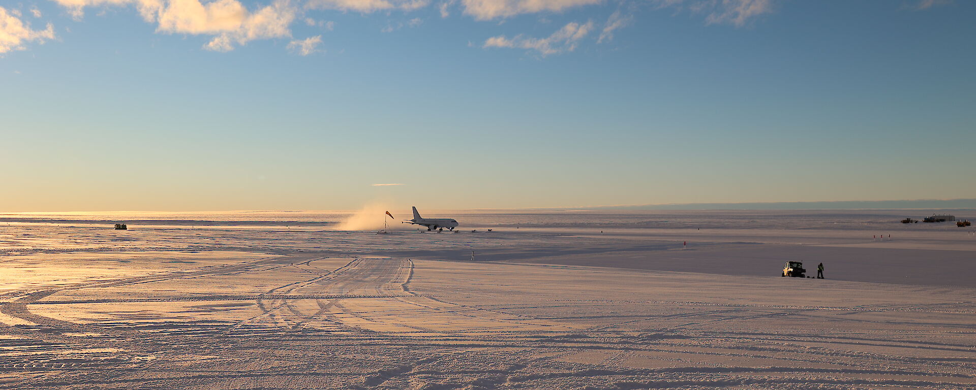 An A319 passenger plane has just landed on a wide, flat plain of snow and ice. A cloud of snow is kicked up in its wake as it goes down the ice runway. The glow of sunrise gently lights the scene