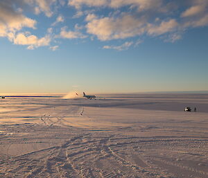 An A319 passenger plane has just landed on a wide, flat plain of snow and ice. A cloud of snow is kicked up in its wake as it goes down the ice runway. The glow of sunrise gently lights the scene