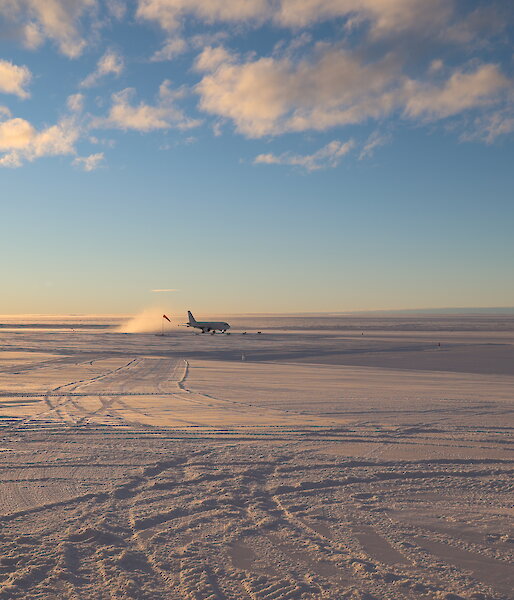 An A319 passenger plane has just landed on a wide, flat plain of snow and ice. A cloud of snow is kicked up in its wake as it goes down the ice runway. The glow of sunrise gently lights the scene