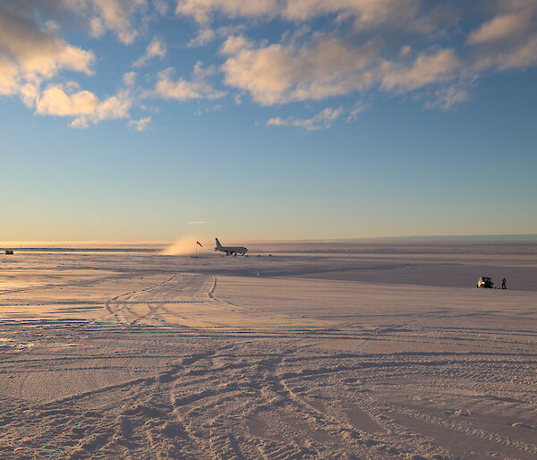 An A319 passenger plane has just landed on a wide, flat plain of snow and ice. A cloud of snow is kicked up in its wake as it goes down the ice runway. The glow of sunrise gently lights the scene