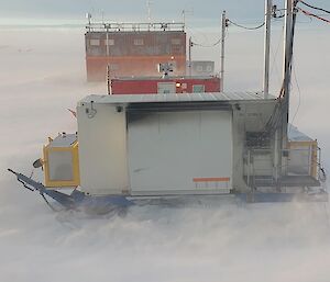 A few small buildings created out of converted shipping containers, lined up close together in a wide, snowy plain. The ground looks soft and featureless, suggesting that a strong wind is blowing much of the snow along the ground, and the sky is full of grey clouds