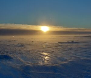 The sun is just rising through a thin layer of cloud on the horizon into a clear sky. The ground is flat, though in the foreground, the bumpy texture and deep blue colour of ice is apparent. Thin plumes of drifting snow soften the view of the icy ground