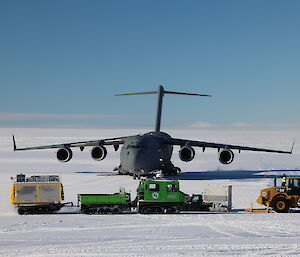 A front view of a dark grey C-17 cargo transport plane parked on flat, white icy ground under a blue sky. In front of the plane are a couple of vehicles to unload the plane's cargo and deliver it to Casey station