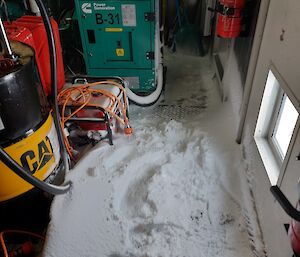 The inside of a plant room, with a generator unit, a few tanks, drums and power cords visible. The room's floor is covered with snow, a few inches thick in some places. Some smaller items close to the floor also have a coat of snow