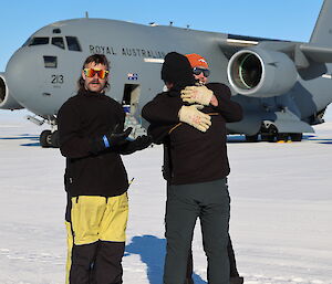 Two men are hugging each other goodbye in front of the C-17 plane. Another man stands beside them making a supplicating gesture, as if wondering if he will get a hug too