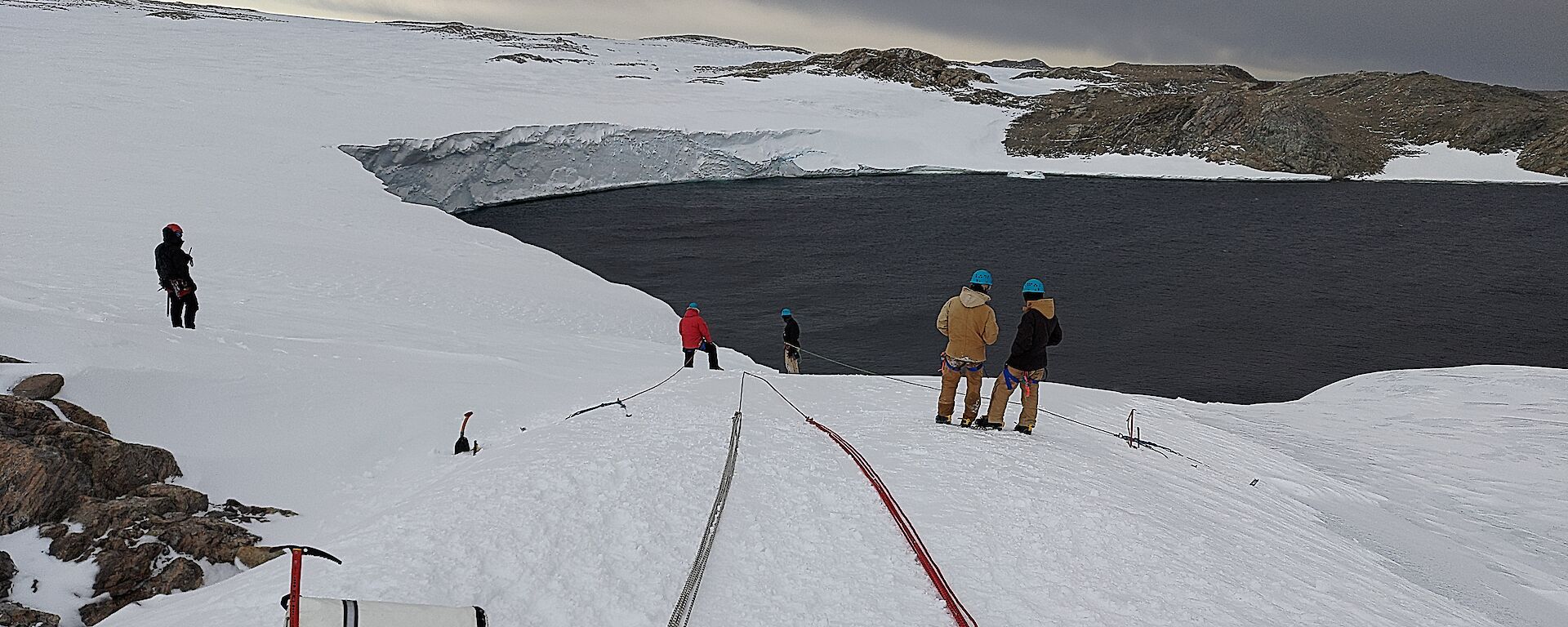 Two long bundles of ropes laid out down a gentle snowy slope, towards an ocean bay. Some people in winter jackets and helmets are inspecting the ropes' layout