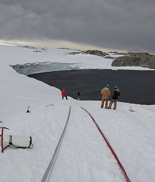 Two long bundles of ropes laid out down a gentle snowy slope, towards an ocean bay. Some people in winter jackets and helmets are inspecting the ropes' layout