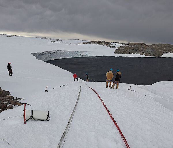Two long bundles of ropes laid out down a gentle snowy slope, towards an ocean bay. Some people in winter jackets and helmets are inspecting the ropes' layout