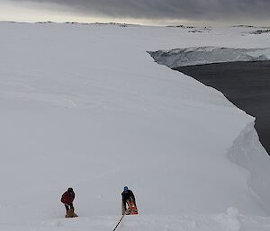 Three people at the bottom of a snowy slope, seen from a distance. One person is lying on the ground as if injured. The second person is bending over the first as if speaking to them. The third person is holding rescue/recovery gear that is attached to a rope coming down the slope