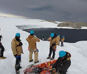A number of people in thick winter clothing and blue safety helmets standing around the top of a snowy slope that leads down to an ocean bay. In the foreground, one of the people is handling some ropes attached to a rescue stretcher