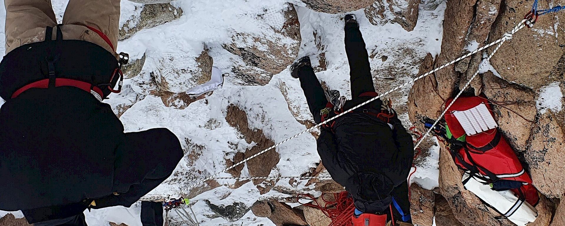 About a dozen people in winter gear and safety helmets have positioned themselves along a downward-sloping gully amidst rocks and snow. A couple of ropes are anchored at the top of the gully: several people are pulling at the rope arrangement, testing its strength