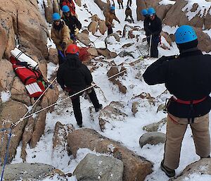 About a dozen people in winter gear and safety helmets have positioned themselves along a downward-sloping gully amidst rocks and snow. A couple of ropes are anchored at the top of the gully: several people are pulling at the rope arrangement, testing its strength