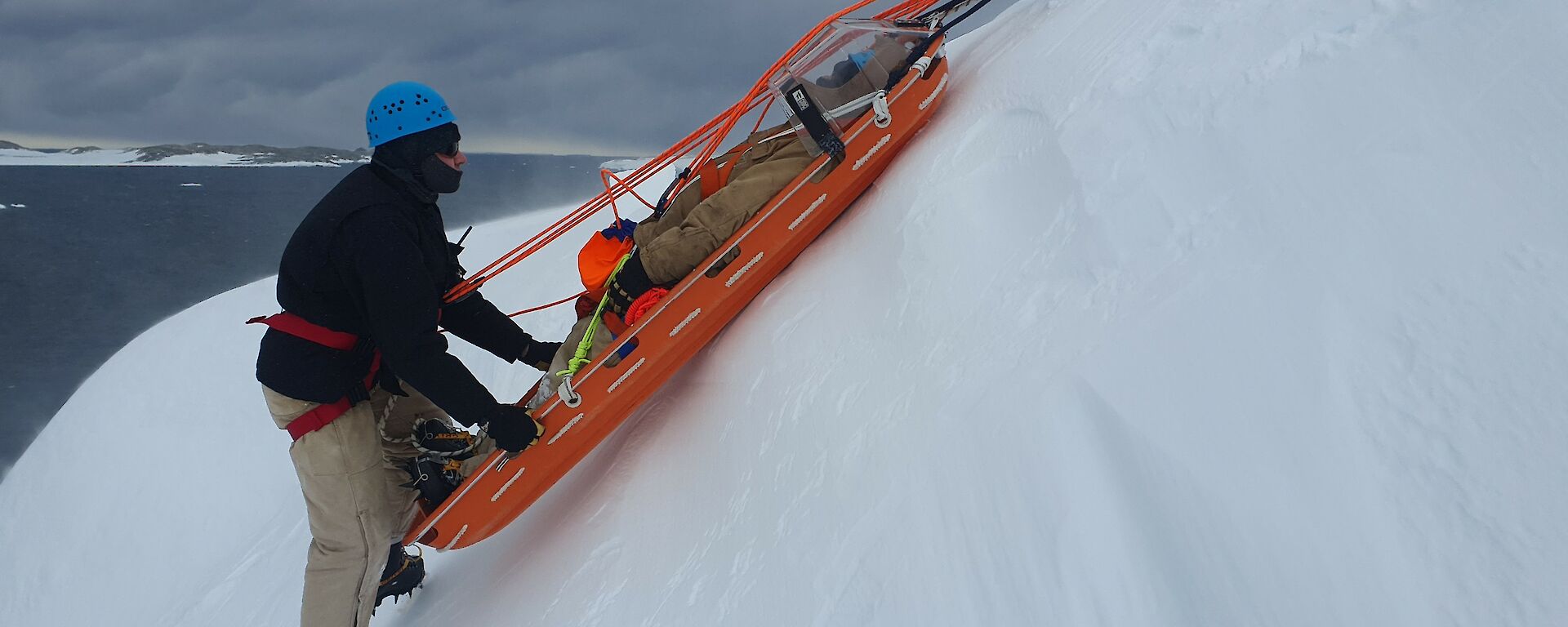 A person in thick winter clothing and a safety helmet is supported on a steep, snowy slope by a harness and ropes. He is holding onto a stretcher with a patient strapped inside. The stretcher has ropes attached to pull it up the slope