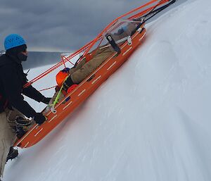 A person in thick winter clothing and a safety helmet is supported on a steep, snowy slope by a harness and ropes. He is holding onto a stretcher with a patient strapped inside. The stretcher has ropes attached to pull it up the slope