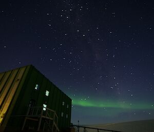 Taken at night. In foreground left is large square building and to right is mound of ice plateau. Sky is clear with bright stars and then across bottom third is green light of Aurora