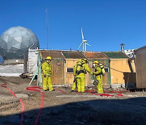 In foreground four people are dressed in fire turn out gear, two wearing breathing aperatus. Behind them is a small wooden hut and then in background is ANARA satellite dome and top of the wind turbine