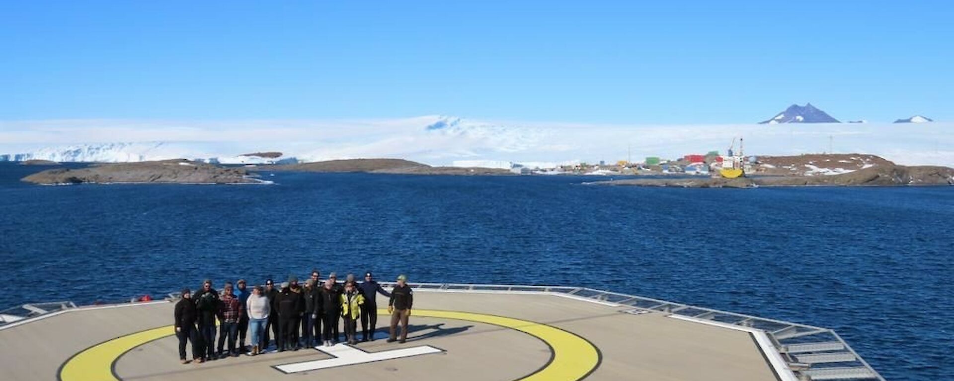 A team of 15 people stand in the middle of a ship's heli-deck with Mawson station in the background
