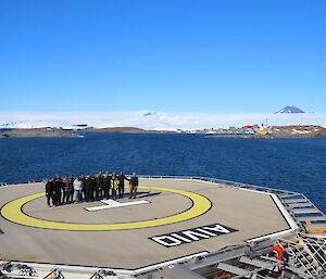 A team of 15 people stand in the middle of a ship's heli-deck with Mawson station in the background