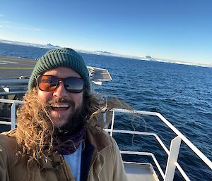 Selfie of man wearing beanie and sunglasses with long hair and beard, smiling with ship railing behind and in the distance the coast of Antarctica