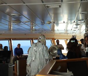 A crowd of people on the bridge of a ship, in the foreground a man dressed as King Neptune with two 'sea-witches' standing to his right