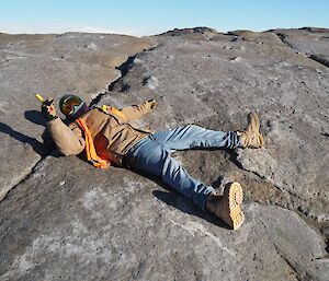 Man in jeans, brown jacket and high vis vest lies on rockground hold aloft an as yet unlit flare