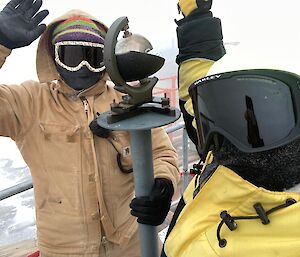Two people rugged up in beanies, goggles, gloves and coats, stand with arms in air either side of a meteorological instrument (sunlight recorder) which is positioned on a metal pole to head heightr)