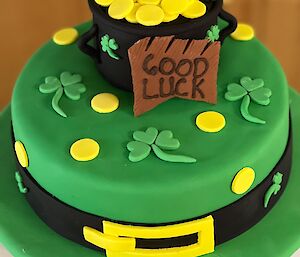 A two teir cake, iced in green, bottom layer has a black belt with gold buckle around it and shamrocks and gold coins on top, top tier is designed to look like a pot of gold, with leprechaun legs sticking out of the gold coins