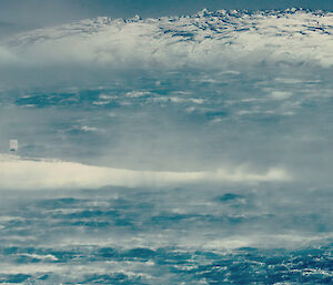 In the foreground and middle is open water with large waves and sea spray blowing, in middle from left is a promitory of land extending into water which is ice covered and at background and rocky island which is partially covered in snow.