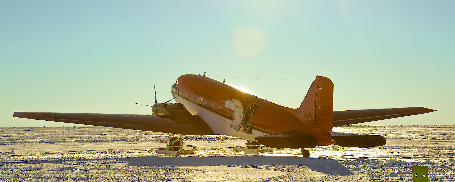 A Basler aircraft, bright orange with a white underside and ski apparatus fixed to its front wheels, is parked on a flat, snowy plain under a clear blue sky