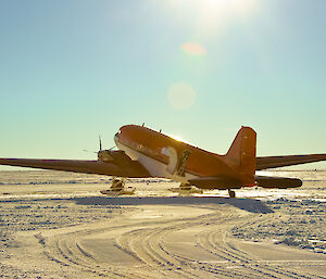 A Basler aircraft, bright orange with a white underside and ski apparatus fixed to its front wheels, is parked on a flat, snowy plain under a clear blue sky