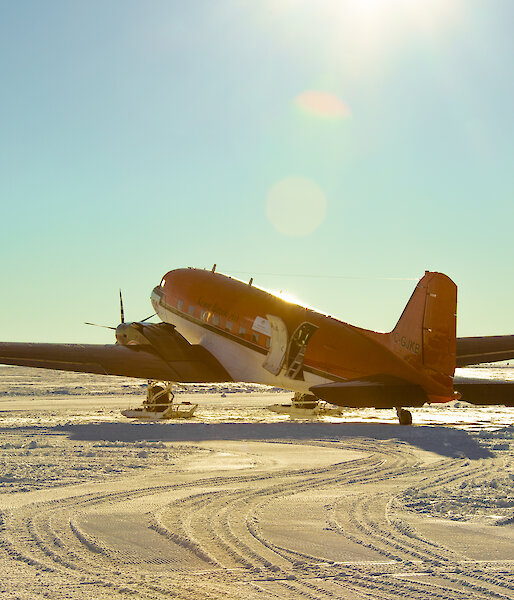 A Basler aircraft, bright orange with a white underside and ski apparatus fixed to its front wheels, is parked on a flat, snowy plain under a clear blue sky