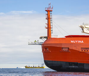 The front end of a large, red icebreaker ship, showing a mast, a crane, and a short walkway extending forwards from the prow. A person in high-vis clothing stands in the centre of the walkway. The name "Nuyina" is painted in white letters on the ship's side. A cargo ship and icebergs can be seen in the distance