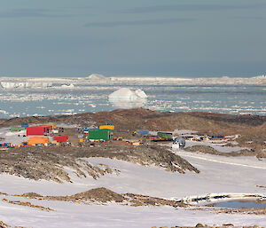 A view of the station, looking down from higher ground at a distance. The station appears as an assortment of rectangular buildings and containers in bright red, yellow, green and blue, spread over a rocky peninsula. In the background, the smooth, pale blue ocean is littered with large and small chunks of ice