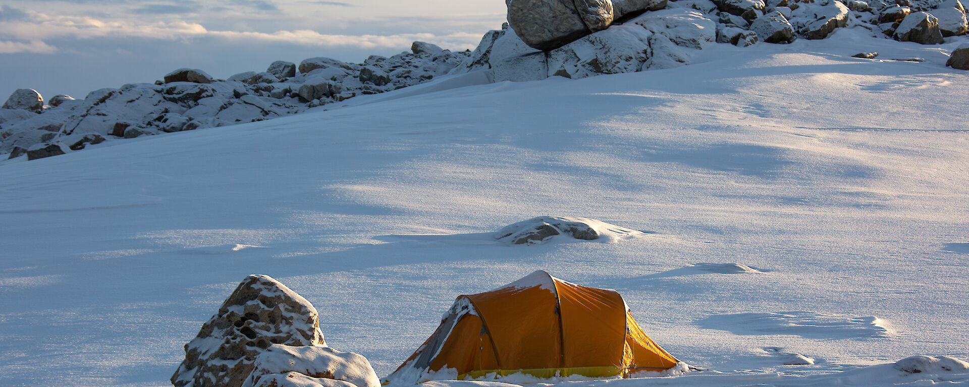 An orange camping tent pitched on smooth, snow-covered ground surrounded by partially snow-covered rocks. Long shadows on the ground suggest it is late in the afternoon