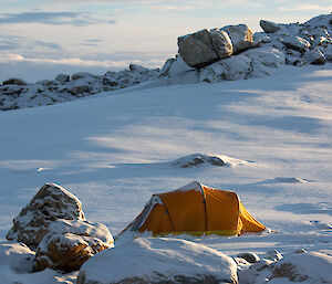 An orange camping tent pitched on smooth, snow-covered ground surrounded by partially snow-covered rocks. Long shadows on the ground suggest it is late in the afternoon
