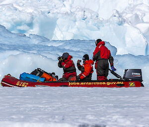 Three people in red and black safety suits are riding in a red inflatable boat driven by an outboard motor. The water is crammed with pack ice and tall, jagged ice formations float nearby, filling the background of the scene. Two of the people in the boat are respectively holding up a camera and a smartphone to take pictures. The third person stands near the motor to steer the boat