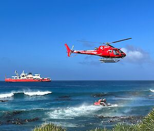 A helicopter hovers over the surf, with a small boat underneath it and a red ship in the background