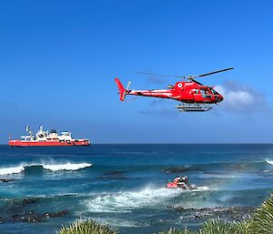 A helicopter hovers over the surf, with a small boat underneath it and a red ship in the background