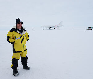 A man in a yellow jacket and beanie standing on the ice with a plane behind him