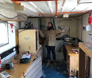 Man stands inside hut which was previously filled with snow with heater standing on floor centre picture. Bench, lab equipment and floor now clear of snow