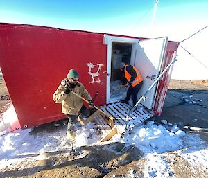 Red square hut with door open, one man standing at door digging out snow from inside the hut while a second man sweeps snow off the door steps