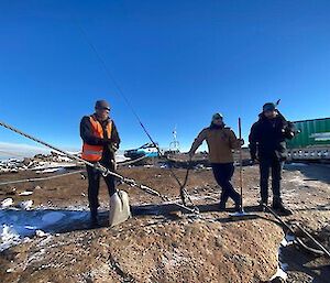 Three men standing on rocky ground holding shovels and wearing winter coats, beanies and gloves. Blue skies above.