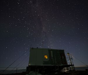 Small square hut in foreground centre of picture with the backdrop of night sky with many bright stars